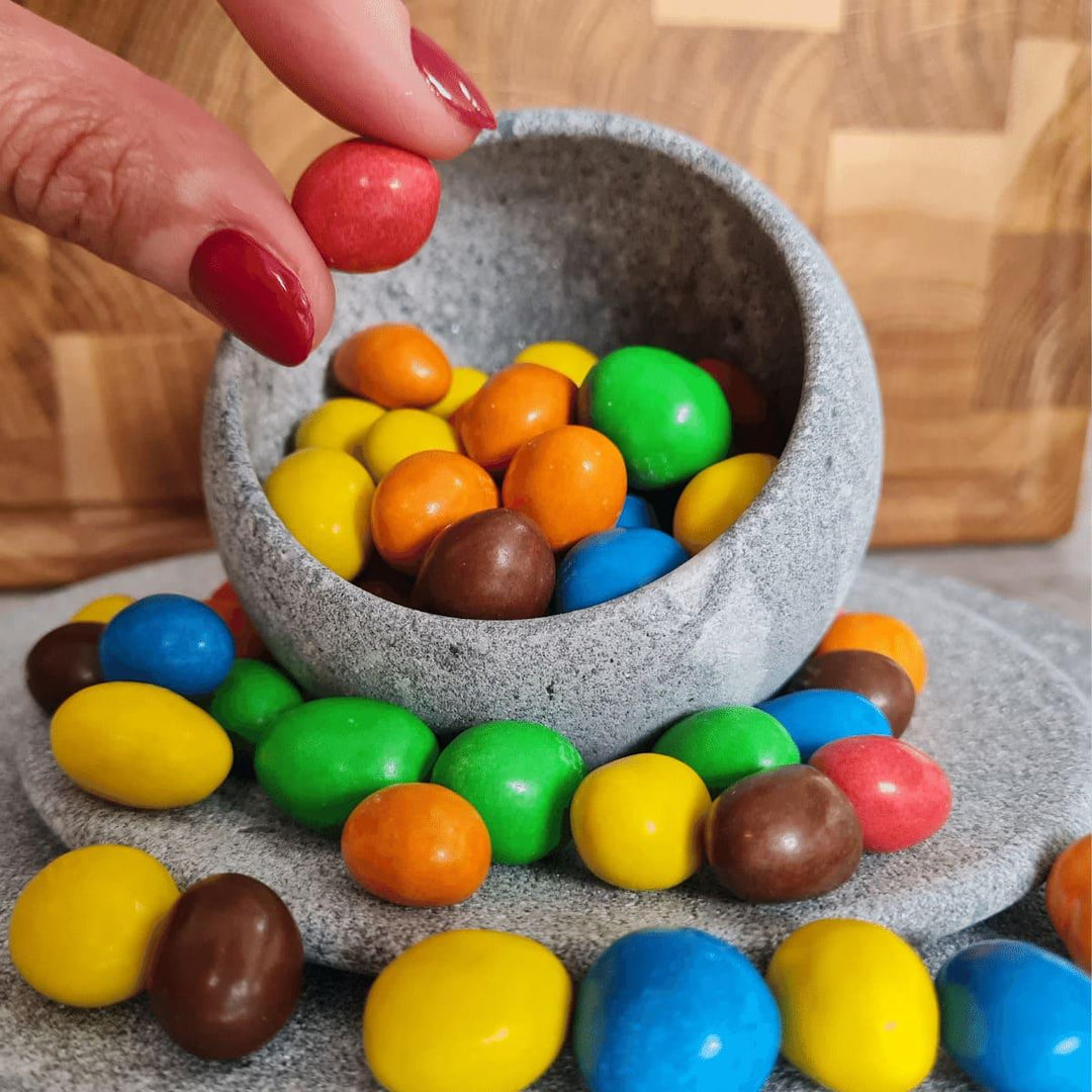 Zwei Finger mit rot lackierten Nägeln greifen ein roten Schokosnack aus einer kleinen, grauen Schale aus Naturstein, die eine schräge Öffnung hat, damit man Snacks und Naschereien besser greifen kann. Rundherum liegen noch weitere, bunte Snacks auf einem grauen Teller aus Naturstein, im Hintergrund ist eine Holzwand.