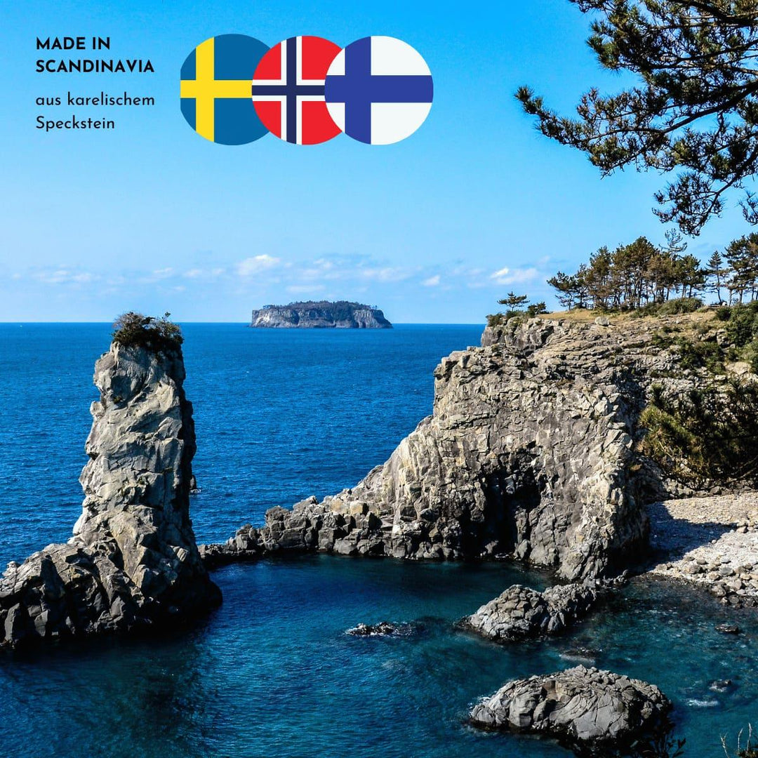 Sonnige Stimmung am Meer mit Felsformationen und Wald am unteren und rechten Bildrand. Drei skandinavische Flaggen und der Hinweis, dass die Produkte aus karelischem Speckstein sind, finden sich im oberen linken Bildrand.