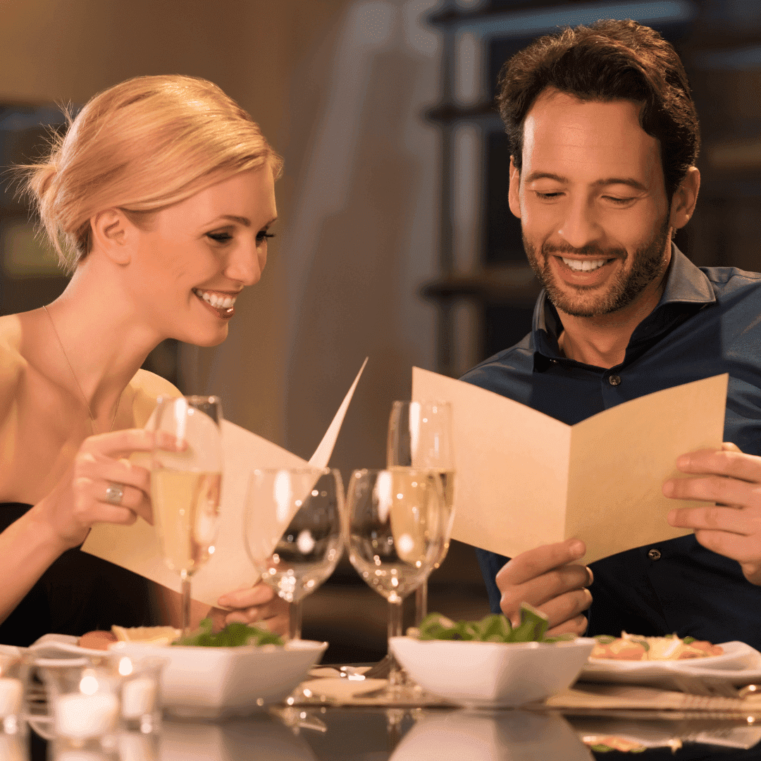 Eine blonde Frau und ein dunkelhaariger Mann mit Bart schauen am schick eingedeckten Restauranttisch in die Speisekarte und lächeln.