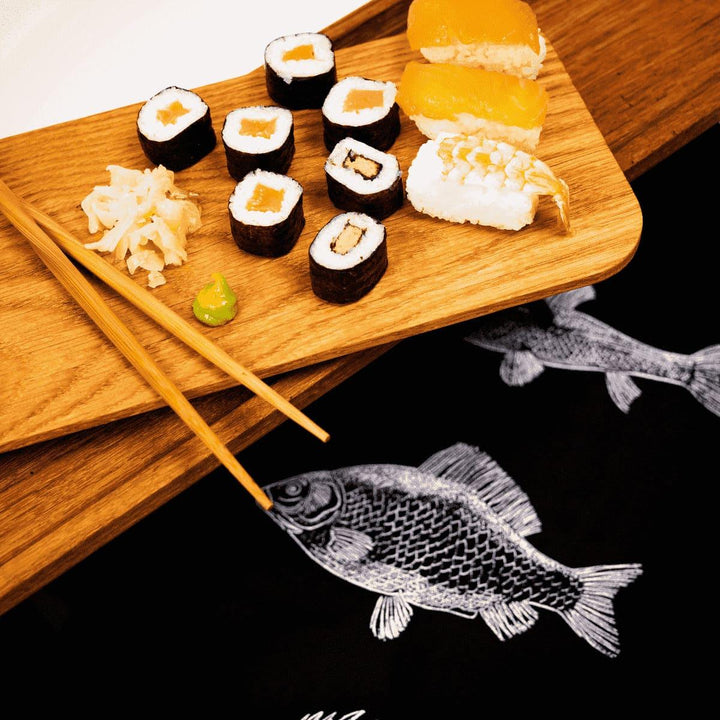 Detailausschnitt einer mit Sushi, Stäbchen und Ingwer dekorierten längliche Etagere aus Eiche Altholz mit schräg drapiertem Tablett auf einer schwarzen Decke mit weißen Fischen