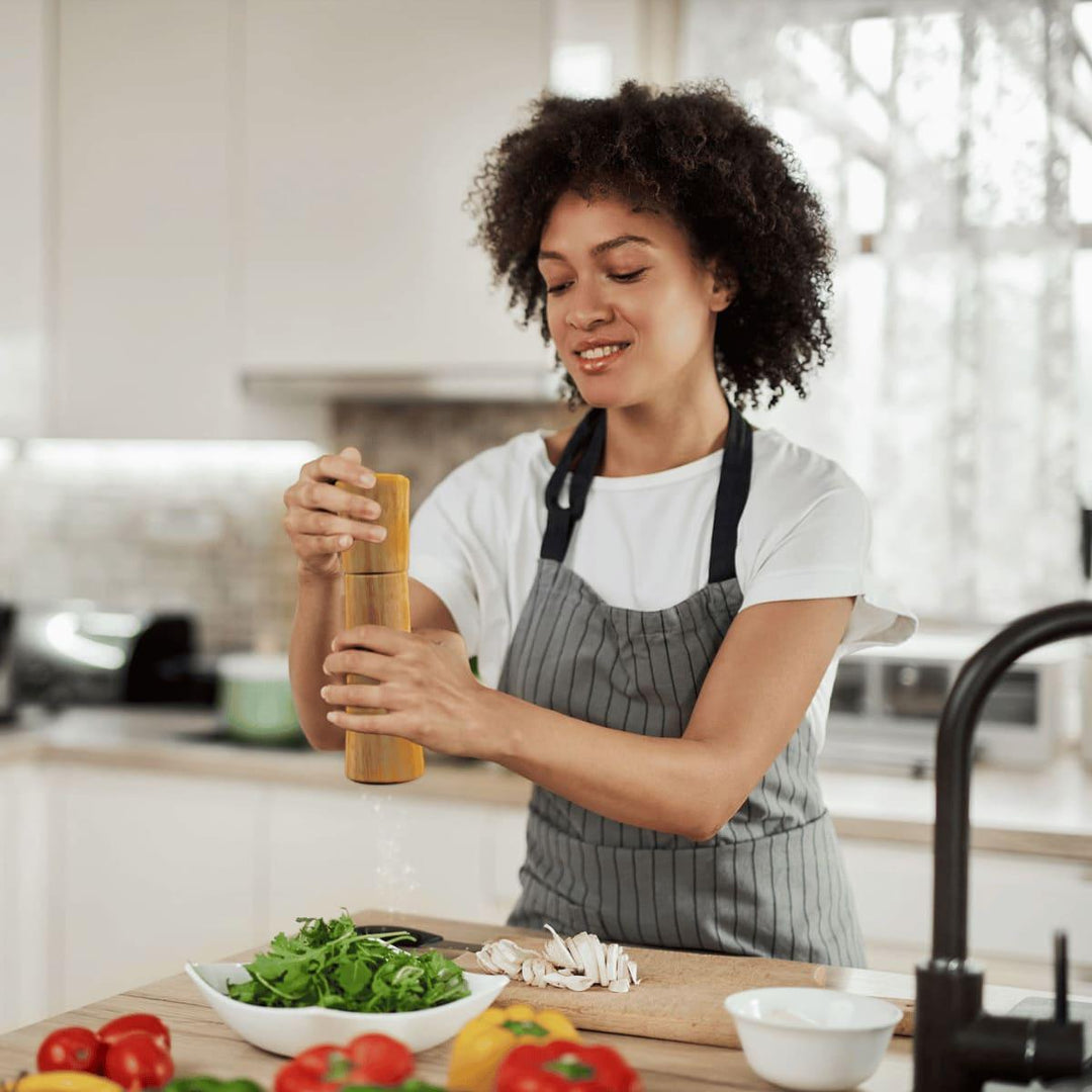 Eine junge, lächelnde Frau mit Schürze würzt in einer hellen Küche ihren grünen Salat mit der Altholz Gewürzmühle PremiumSpice.