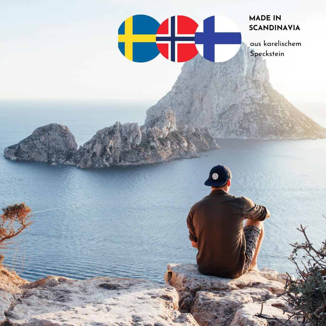 Auf dem Bild sitzt ein junger Mann auf Steinfelsen mit dem Blick aufs Meer und eine Felsformation. Drei skandinavische Flaggen und der Hinweis, dass die Produkte aus karelischem Speckstein sind, finden sich im oberen rechten Bildrand.