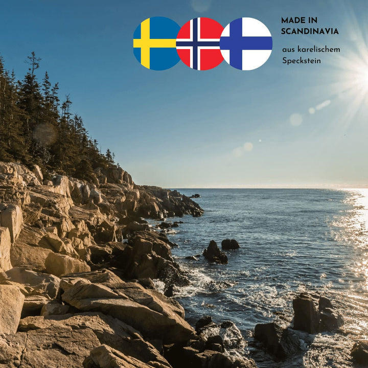Sonnige Stimmung am Meer mit Felsformationen und Wald am linken Bildrand. Drei skandinavische Flaggen und der Hinweis, dass die Produkte aus karelischem Speckstein sind, finden sich im oberen rechten Bildrand.