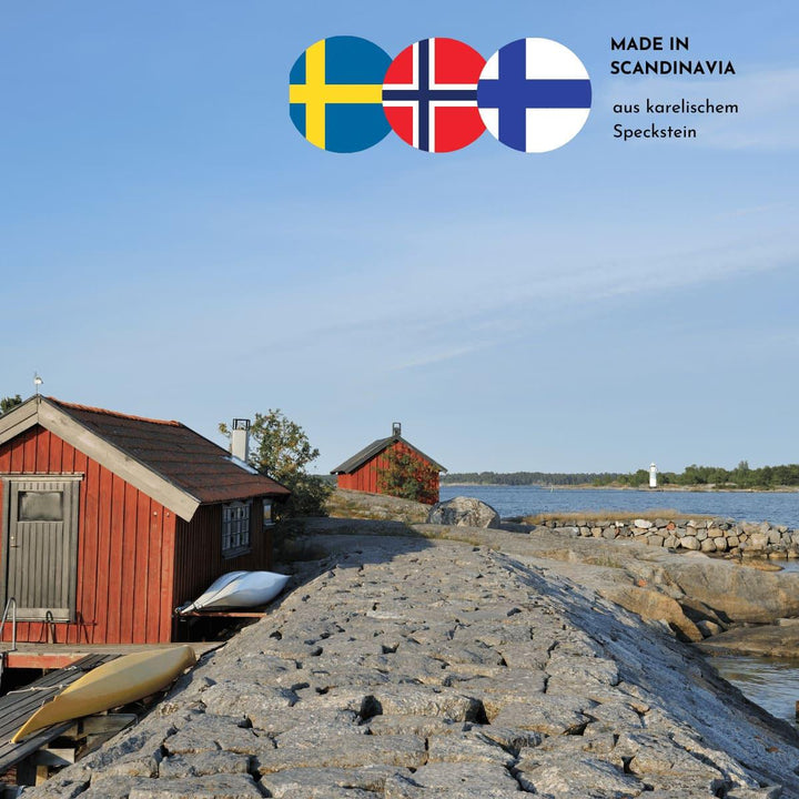 Eine Steindüne mit kleinen roten Holzhäusern mit angelehnten Kanus und der Blick auf das Wasser im Hintergrund zeigen ein skandinavisches Ambiente. Drei skandinavische Flaggen und der Hinweis, dass die Produkte aus karelischem Speckstein sind, finden sich im oberen rechten Bildrand.