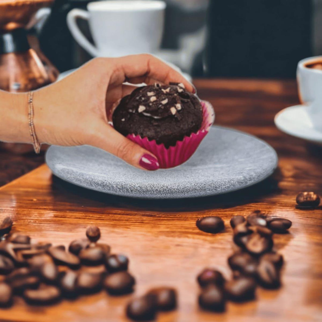 Ein grauer Teller aus Naturstein, karelischer Steatit aus Skandinavien. Eine Hand nimmt sich einen Muffin vom Teller, der auf einer Holztischplatte steht auf dem Kaffeebohnen liegen und Kaffeetassen stehen.