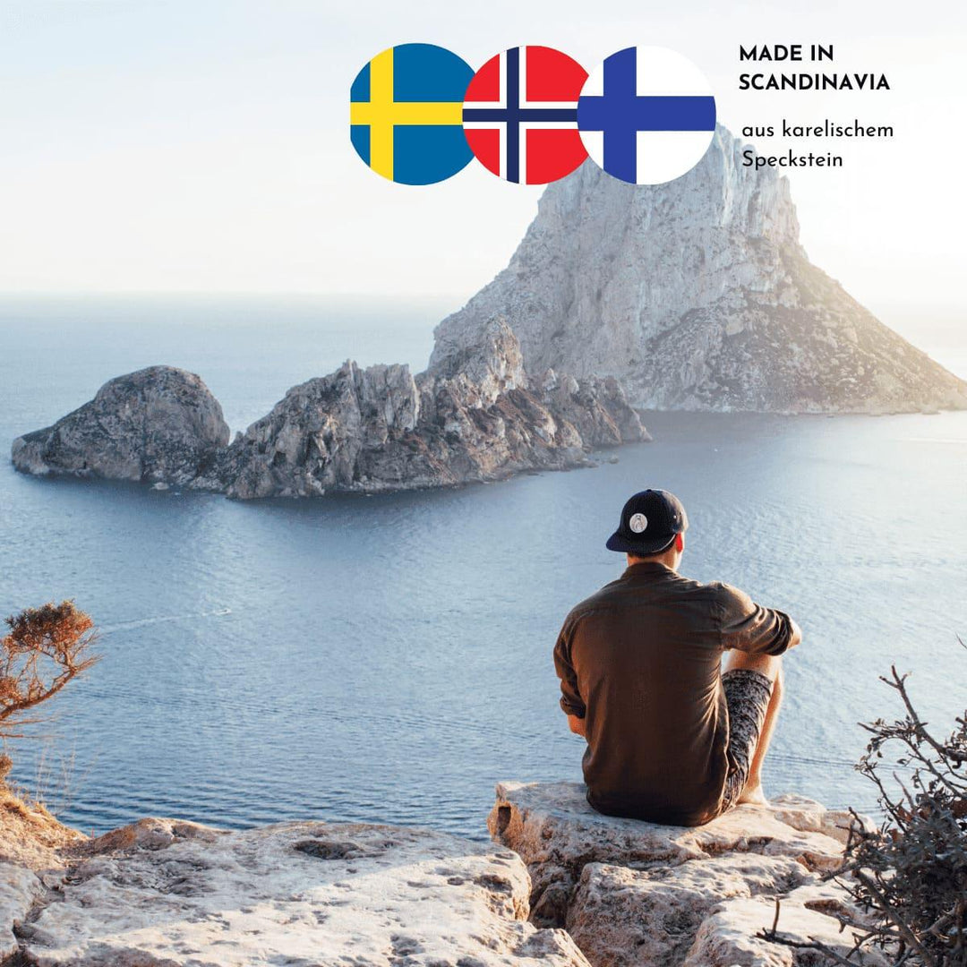 Ein junger Mann mit Cap sitzt am Rand einer Steinküste und blickt über das Meer und auf Felsformationen. Drei skandinavische Flaggen und der Hinweis, dass die Produkte aus karelischem Speckstein sind, finden sich im oberen rechten Bildrand.