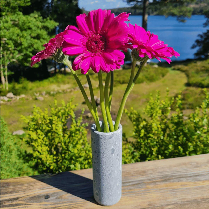 Eine graue, schmale Vase aus Naturstein, karelischer Steatit aus Skandinavien, in der pinkfarbene Blumen stehen. Im Hintergrund ist ein sonniger, grüner Garten am See.