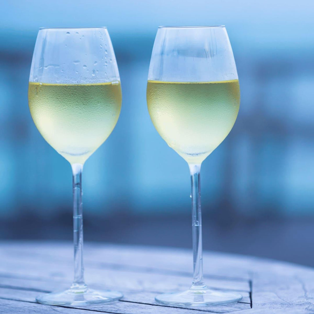 Zwei mit Weißwein gefüllte Weingläser mit kondensiertem Wasser am Glas, die auf einem Holztisch stehen, der Hintergrund ist kühl, bläulich.
