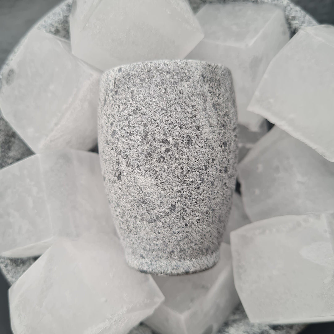 Ein kleines graues Schnapsglas aus Naturstein, karelischer Steatit aus Skandinavien, aus denen man besonders gut klaren, kalten Schnaps, signalisiert durch die Eiswürfel auf dem das Glas liegt, trinken kann.