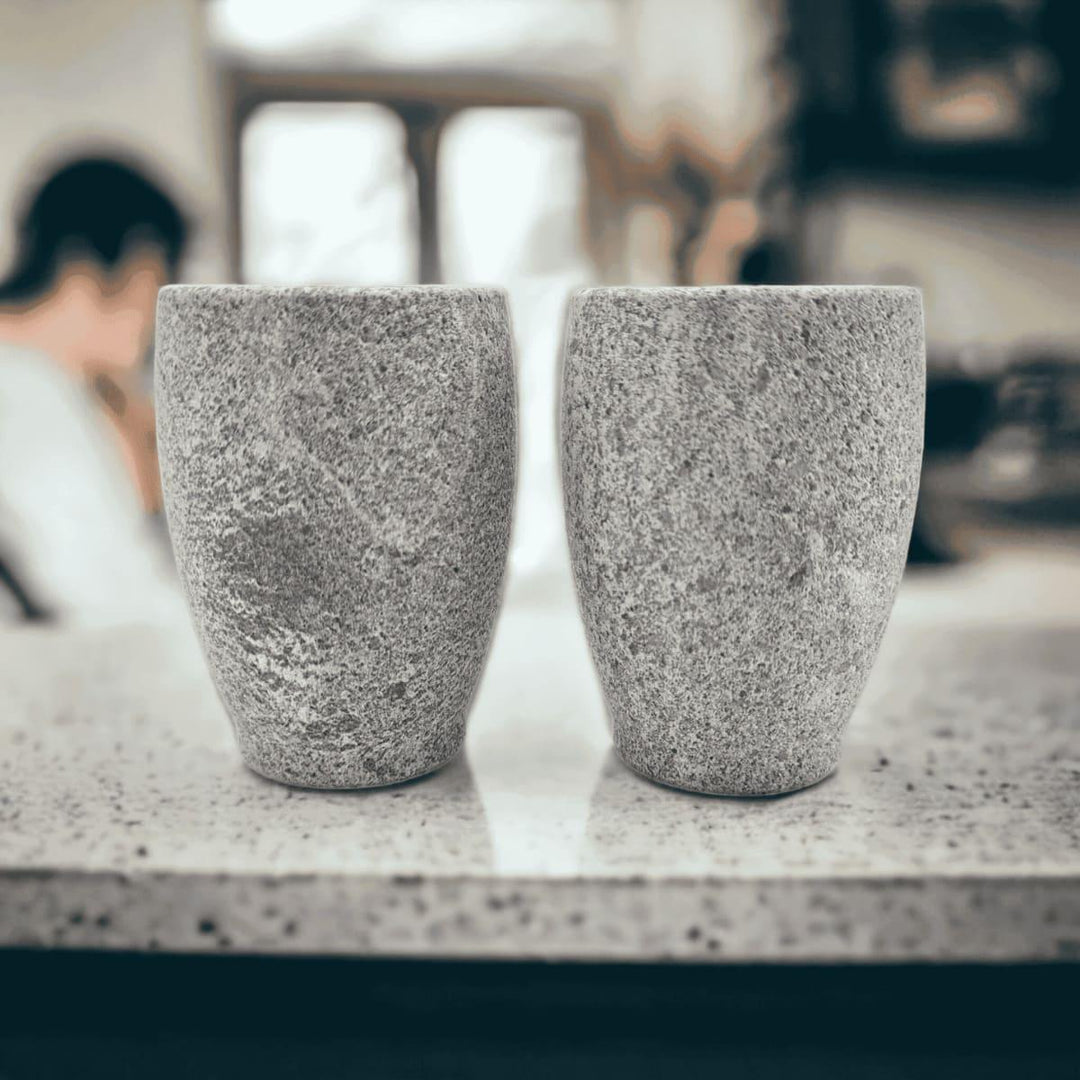 Zwei kleine graue Schnapsgläser aus Naturstein, karelischer Steatit aus Skandinavien, stehen auf einer Arbeitsplatte aus Granit in einer Küche, im Hintergrund verschwommen steht ein Mensch.
