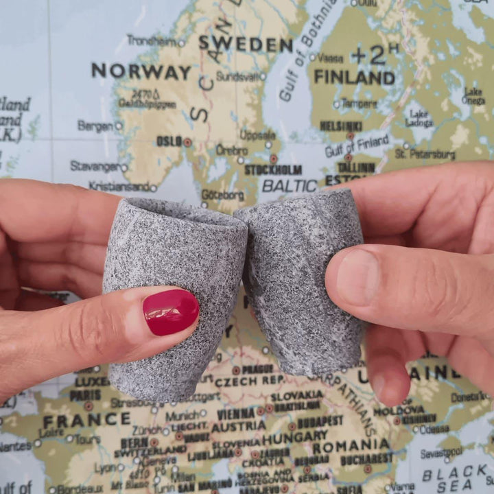 Eine weibliche und eine männliche Hand prosten sich mit zwei kleinen grauen Schnapsgläsern aus Naturstein, karelischer Steatit aus Skandinavien, vor einer Weltkarte auf der man Skandinavien sieht, an.