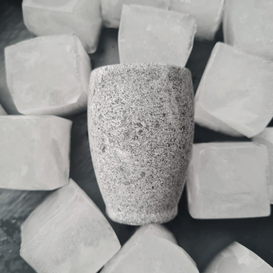 Ein kleines graues Schnapsglas aus Naturstein, karelischer Steatit aus Skandinavien, aus denen man besonders gut klaren, kalten Schnaps, signalisiert durch die Eiswürfel auf dem das Glas liegt, trinken kann.